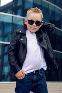 Baby boy in sunglasses and black leather jacket smiling on the background of the glass building