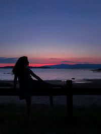 Silhouette woman sitting by sea against sky during sunset