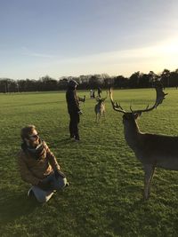 Man sitting by stag on grassy field during sunset