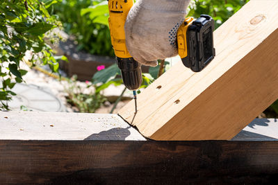 A carpenter works with a yellow electric screwdriver, twists a screw into a wooden plank beam