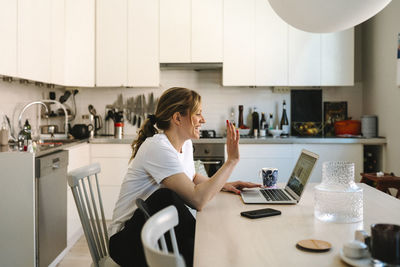 Happy female freelancer waving on video call through laptop at table in kitchen