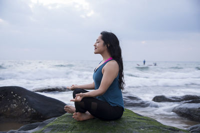 Side view of woman meditating on rock by sea against sky