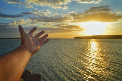 Cropped hand gesturing over sea against sky during sunset