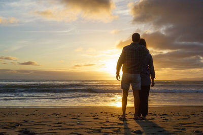 Rear view of man standing on beach against sky during sunset