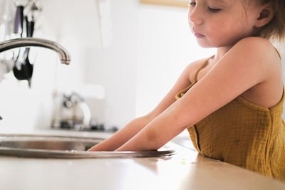 European child of four years old in yellow jumpsuit washes his hands in real bright kitchen