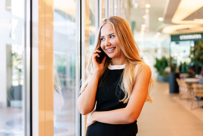 Cheerful female manager talking on cellphone and looking away with smile while standing near window of modern mall in daytime