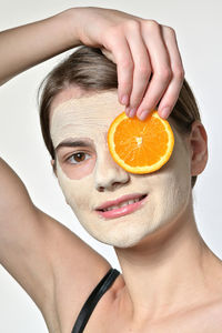 Portrait of woman with facial mask holding orange slice against white background