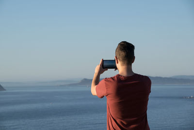 Rear view of man photographing sea against clear sky