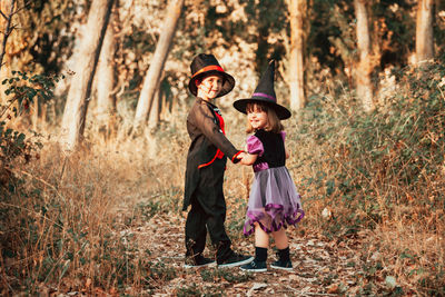 Full length portrait of girl wearing witch hat standing with brother at forest during halloween