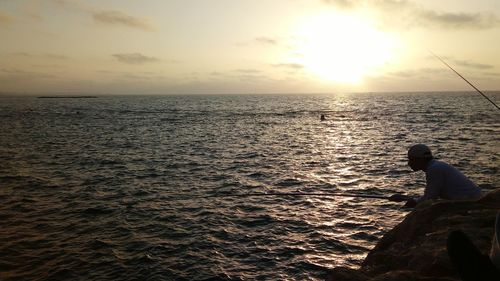 Rear view of silhouette man fishing in sea against sunset sky