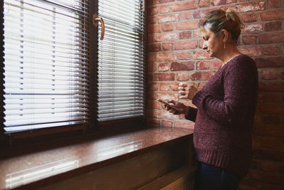 Woman texting on smartphone, drinking coffee while taking a break in office standing at window