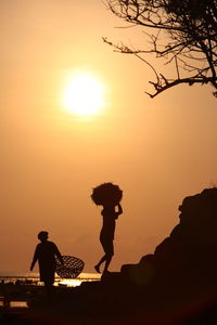 Silhouette worker carrying basket while walking on rock against orange sky