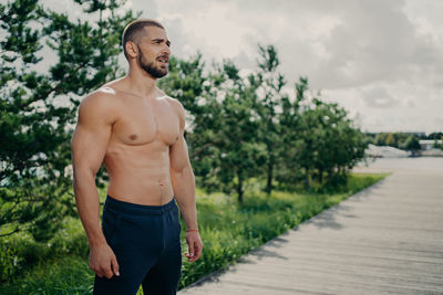 Young shirtless man looking away while standing against plants