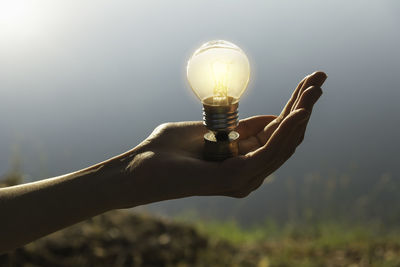 Close-up of hand holding light bulb against sky