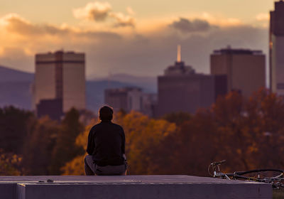 Rear view of man sitting, watching sunset against city skyline 