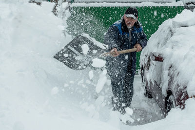 A man is shoveling snow from the area near the house. nearby is a car covered with snow.