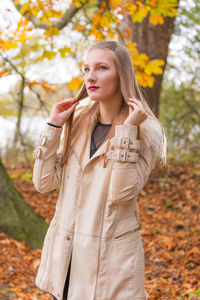Young woman touching her blond hair while standing by trees during autumn