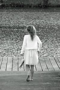Rear view of girl holding shoes standing on pier over lake
