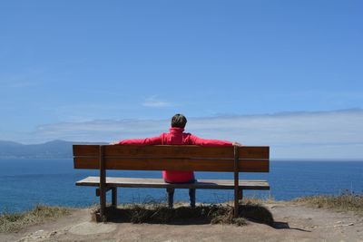 Rear view of man sitting on bench at beach against blue sky