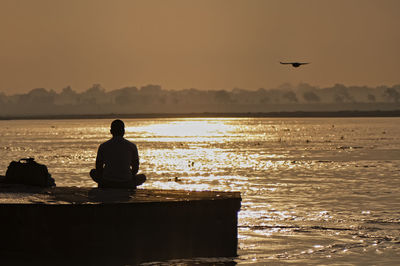 Rear view of silhouette man sitting on beach against sky during sunset