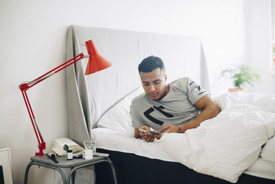 Mid adult man holding glaucometer and mobile phone while lying on bed by side table