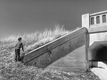 Rear view of boy standing by built structure against sky during sunny day