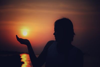 Optical illusion of silhouette woman holding sun while standing against sea during sunset