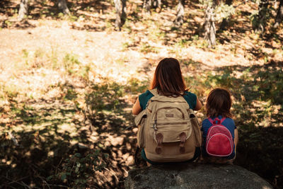 Rear view of mother and daughter with backpacks sitting on rocks in forest