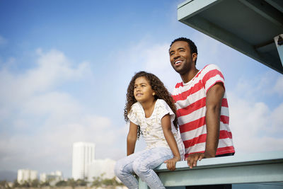 Father and daughter looking away while relaxing at railing on pier against sky
