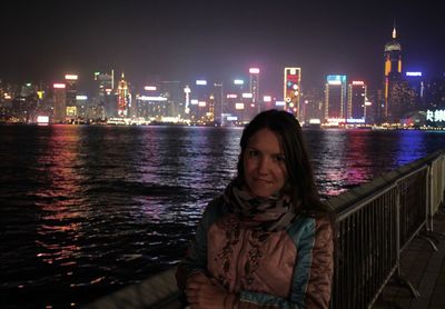 Portrait of woman standing by river in illuminated city at night