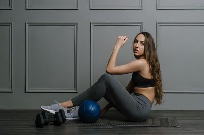 Portrait of young woman sitting by medicine ball against wall
