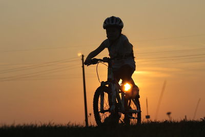 Silhouette boy riding bicycle on field against sky during sunset