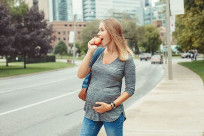 Pregnant woman eating apple while standing on sidewalk in city