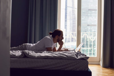 Man in a white t-shirt, a freelancer with glasses, lie on the bed at home with a laptop and works 