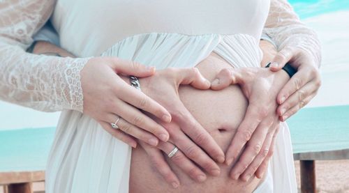 Cropped hands of man making heart shape on pregnant wife abdomen at beach