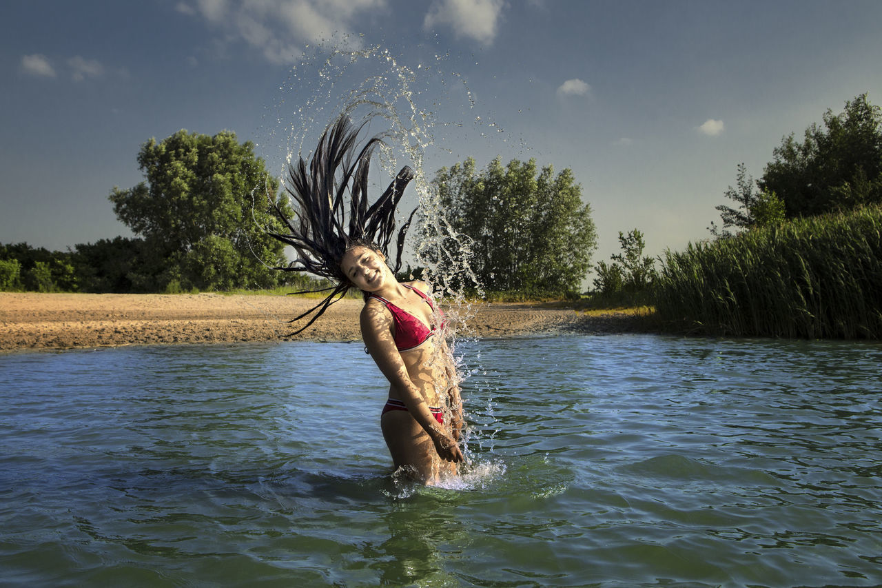 Girl splashing water while tossing hair in lake against sky