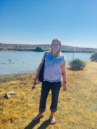 Portrait of smiling mid adult woman standing on field by lake against clear sky during sunny day