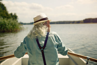 Back view of senior man sitting in rowing boat on a lake wearing suspenders and summer hat