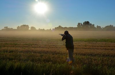 Rear view of man aiming gun while standing on grass against sky