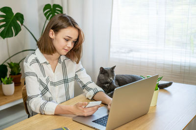 Young woman entrepreneur working on laptop at home with her cat, together in modern room with plants