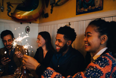 Happy multi-ethnic male and female friends holding burning sparklers in restaurant during dinner party