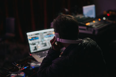Rear view of dj using laptop and sound mixer while wearing headphones at night