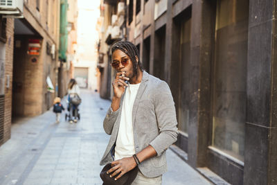 Portrait of young man wearing sunglasses smoking while standing at alley against building in city