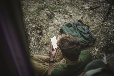 High angle view of man sitting cross-legged while using smart phone against motor home during camping
