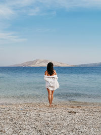 Rear view lifestyle image of stylish young woman in white shirt standing on beach and looking at sea