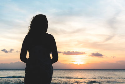 Rear view of silhouette woman standing at beach against sky during sunset