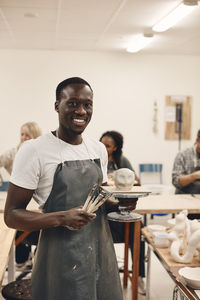 Portrait of smiling young man with work tools and molded clay standing in art class