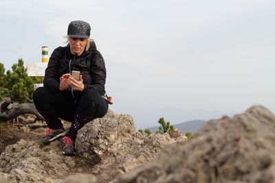 Full length of mature woman using mobile phone while sitting on rocks against sky