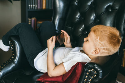 The red-haired boy cuts in an armchair and holds a phone in his hands.