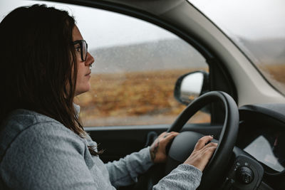 Side view of young woman in eyeglasses with piercing driving automobile between wild lands with hills in rainy weather